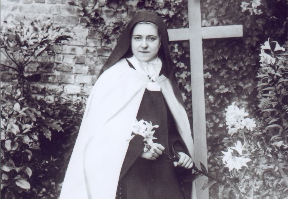 St. Therese holding lily, in front of cross.